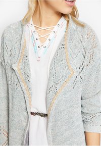 Vaaleansininen neulecardigan, jossa on pitsikuvioita, aallotetut reunat ja herkkä trimmaus, käytetty valkoisen, pitsiyksityiskohdalla varustetun topin päällä.