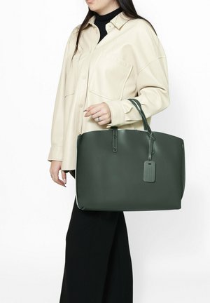 Borsa tote in pelle verde con due manici, forma rettangolare, superficie liscia e un'etichetta attaccata. Indossata da una persona con una camicia chiara.