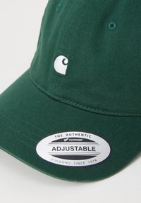 Cappellino verde regolabile realizzato in tessuto testurizzato, con logo ricamato bianco e un'etichetta circolare che ne attesta l'autenticità.