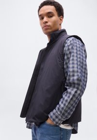 Νέος άνδρας με τα χέρια στις τσέπες, φοράει μαύρο αμάνικο γιλέκο πάνω από ένα μπλε πουκαμισάκι με καρό μοτίβο και μπλε τζην, σε απλό λευκό φόντο.