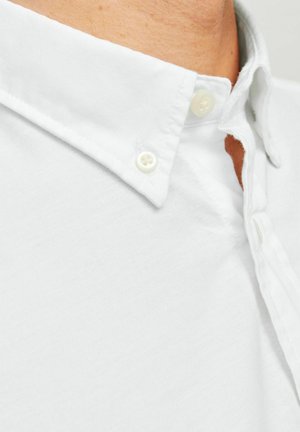 Chemise blanche à boutons avec un col classique, en tissu de coton. Présente un tissage texturé et des boutons en plastique au col et sous la patte de boutonnage.