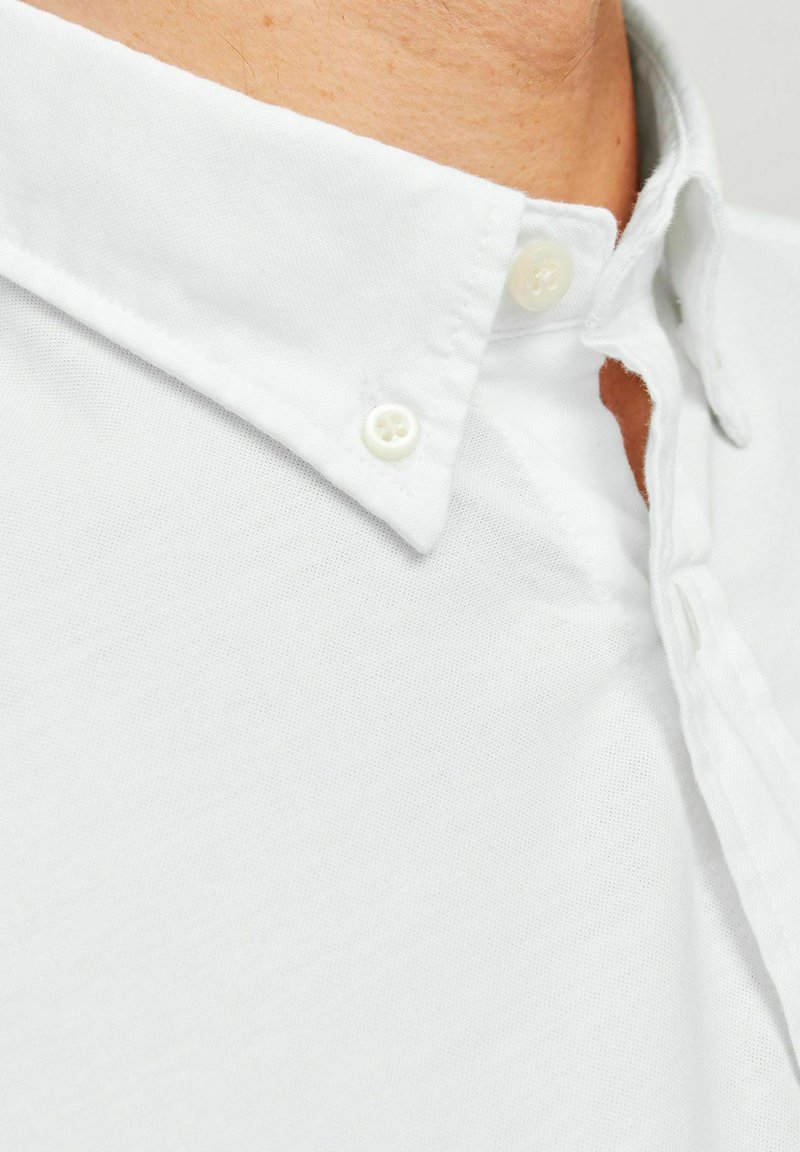 Chemise blanche à boutons avec un col classique, en tissu de coton. Présente un tissage texturé et des boutons en plastique au col et sous la patte de boutonnage.