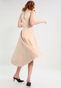 Beige ärmlös midiklänning med en åtsittande kropp och flödande kjol. Tyget har en slät textur. Bärs med svarta lackskor.
