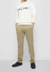 Cremefarbener Sweatshirt mit schwarzem "Tommy Hilfiger"-Logo, kombiniert mit khakifarbenen Chinos und marineblauen Slip-On-Schuhen, präsentiert ein lässiges Outfit.