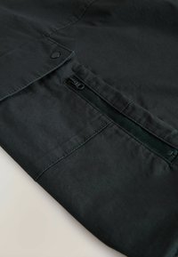 Schwarzes Stoffelement mit einer Klappen-Tasche und seitlichem Reißverschluss. Glatte Textur mit sichtbaren Nähten entlang der Kanten. Sichere Beschläge.