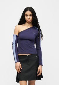 Γυναίκα με μακριά σκούρα μαλλιά που φοράει μωβ μπλούζα χωρίς ώμους με ανοιχτό μπλε ρίγες και μαύρη φούστα με φερμουάρ στο μπροστινό μέρος.