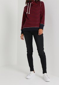 Felpa a righe rossa e blu con collo a imbuto e lacci, abbinata a jeans skinny neri e sneakers bianche.