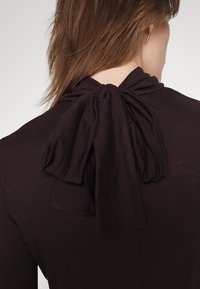 Femme aux cheveux châtain moyen portant un haut foncé avec un grand nœud ample noué à l'arrière du cou, assorti d'une seule boucle d'oreille en or.