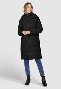 Longue doudoune noire avec capuche, fermeture éclair sur le devant et détails à pression. Associée à un jean bleu et des bottines noires.