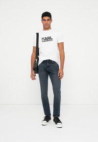 Jaunas vyras stovi, apsirengęs balta Karl Lagerfeld marškinėliais, tamsiai mėlynomis džinsomis, juodais sportbačiais ir nešdamas juodą per petį krepšį ant baltos fono.