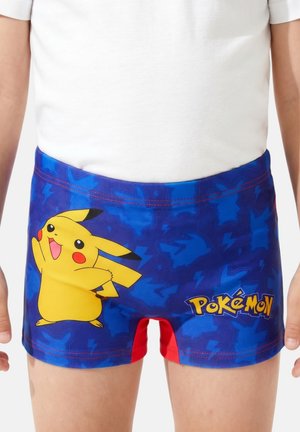 Enfant portant un short de bain bleu avec le personnage Pikachu et le logo Pokémon, debout sur un fond neutre.