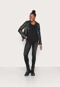 Jqueta de moto de couro preto sobre uma blusa preta, emparelhada com jeans skinny pretos e botas grossas pretas com fivela. Fundo branco.