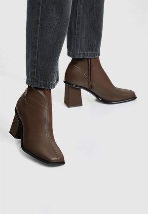 Brown leather ankle boots with a pointed toe, square heel, and side zipper detail, set against dark denim pants.