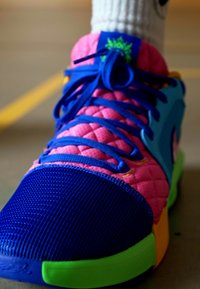 Scarpa atletica colorata con parte superiore in rete blu, sezioni imbottite rosa, suola verde e lacci blu ben visibili. Dettaglio di branding notevole presente.