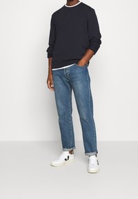 Homme portant un pull marine foncé, un jean bleu retroussé et des baskets blanches avec un logo V noir, debout contre un mur blanc.