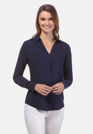Blusa a maniche lunghe blu navy con scollatura a V, dettaglio con piega frontale e polsini con bottoni, abbinata a pantaloni bianchi. Tessuto morbido.