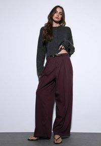 Cardigan gris court à boutons, associé à un pantalon taille haute plissé couleur bordeaux et des tongs noires ; textures lisses, design décontracté.