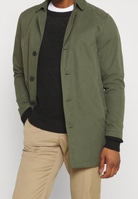 Olivgrön trenchcoat med krage, svarta knappar och sidofickor, lager på lager över en texturerad svart tröja och beige byxor.