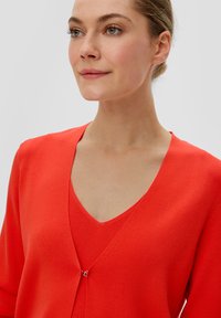 Cardigan en tricot rouge avec un décolleté en V, doté d'une petite fermeture à clip en argent et de manches courtes. Texture lisse, design léger.