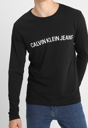 Persona vistiendo una camiseta negra de manga larga con el texto inclinado en blanco "CALVIN KLEIN JEANS", combinada con jeans azules.