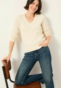 Cremfarbener Strickpullover mit V-Ausschnitt und strukturiertem Zickzack-Muster, kombiniert mit enganliegenden blauen Jeans. Das Model lehnt an einem Holzstuhl.