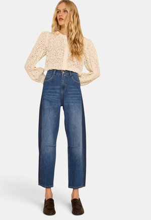 Femme portant un chemisier en dentelle crème à manches longues, un jean bleu taille haute avec des panneaux latéraux foncés, et des mocassins marron foncé, debout les mains sur les hanches.