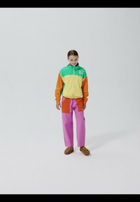 Fille debout portant une veste verte, jaune et orange, un pantalon rose et des chaussures marron, contre un fond uni gris clair.