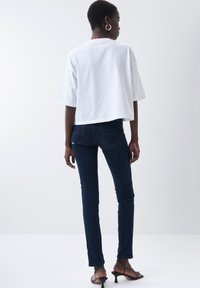 T-shirt blanc court, à manches courtes, associé à un jean slim bleu marine avec poches arrières. Le modèle porte des sandales noires à lanières.