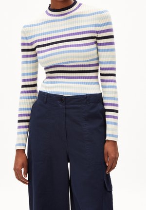 Personne portant un pull à manches longues côtelé avec des rayures horizontales blanches, bleues, violettes et noires, associé à un pantalon taille haute bleu marine.