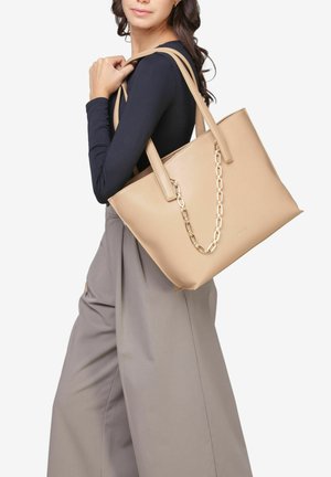 Sac à main en cuir beige avec deux longues poignées et un accent en chaîne dorée. Texture lisse et design structuré. Le modèle porte un pantalon gris.