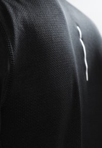 Czarna sportowa koszulka wykonana z teksturowanej dzianiny, z gładkim białym akcentem na ramieniu. Zbliżenie ukazuje szczegóły tkaniny.