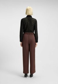 Persoon met kort blond haar, gekleed in een zwart patroon lange mouw blouse en hoge taille bruine wijde broek, staande met de rug naar de kijker gekeerd.