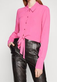 Blusa corta fucsia con chiusura a bottoni e dettaglio con fiocco in vita; maniche lunghe e tessuto leggero e liscio. Pantaloni neri a vita alta.