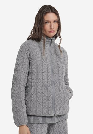 Kvinde iført en grå kabelstrikket lynlåstrøje og matchende bukser med hænderne i lommerne, stående foran en hvid baggrund.