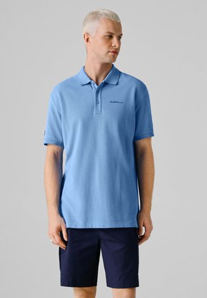 Uomo con capelli biondi corti che indossa una polo azzurro chiaro con la scritta "Red Bull Racing" e pantaloncini blu scuro, guarda verso destra.