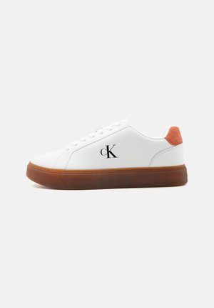 Biały niski sneaker z brązową podeszwą, pomarańczową zawieszką na pięcie, sznurowadłami i logo "cK" po boku.