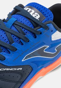 Zapatilla deportiva azul con parte superior de malla, cordones azul oscuro, detalles de ventilación y suela de goma naranja. La marca es visible en la lengüeta.