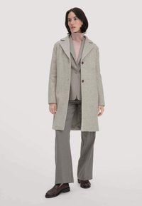 Hellgrauer Wollmantel mit einem Reverskragen und braunen Knöpfen, getragen über einem hellrosa Rollkragenpullover und einem grauen Anzug, kombiniert mit braunen Slippern.