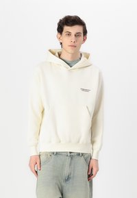 Sudadera con capucha crema con bolsillo delantero, que presenta un pequeño logo bordado a la izquierda. Textura suave y corte relajado. Combinada con jeans claros.