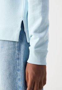 Sudadera de algodón azul claro con puños acanalados y aberturas laterales. Lleva puesta sobre un vaquero azul claro, mostrando un detalle de costura y una textura suave.