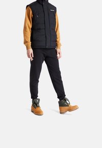 Czarna kamizelka puchowa na brązowej bluzie, w połączeniu z czarnymi spodniami i jasnobrązowymi butami z zielonymi akcentami. Kamizelka ma zamki błyskawiczne.