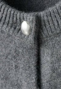 Primer plano de un suéter gris de punto que presenta un cuello acanalado y un gran botón plateado brillante, destacando la suave textura y los detalles.