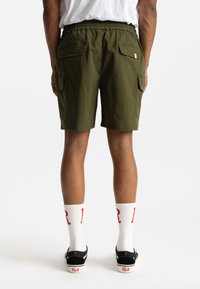 Olijfgroene shorts met een elastische tailleband en twee achterzakken, in combinatie met witte sokken met rode letters en zwarte sneakers.