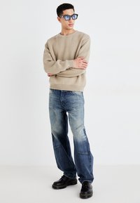 Beige strikket sweater med ribbede manchetter, blå denim jeans med varierende vask, og sorte snørebåndssko med gule stiklinger.