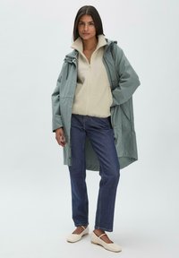 Helle türkisfarbene, wasserdichte Jacke über einem cremefarbenen Fleecepullover, blaue Jeans und beige Schuhe. Die Jacke hat eine Kapuze und Fronttaschen.