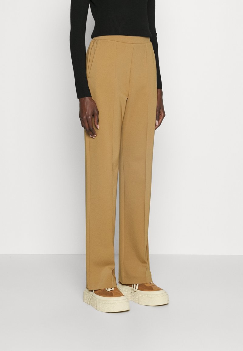 Personne portant un pantalon droit couleur camel, un haut noir à manches longues et des baskets plateformes beiges, debout devant un fond clair uni.