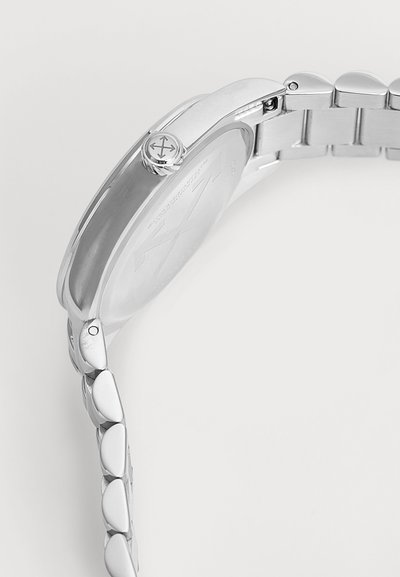 Montre-bracelet en métal argenté vue de côté montrant la couronne avec un logo et un bracelet à maillons sur un fond uni.