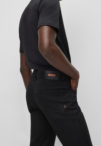 Jeans negros con un parche del logo BOSS en naranja en la cintura, de diseño ajustado y con bolsillos traseros. Tejido liso y líneas limpias.
