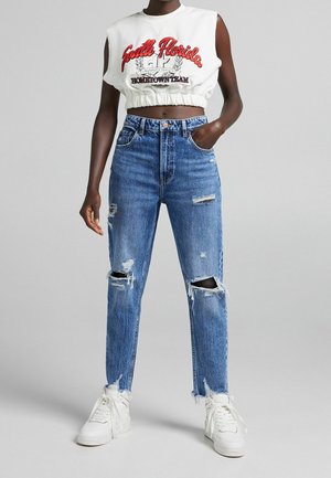 Bílý crop top s červeným nápisem, v kombinaci s vysokými, roztrhanými modrými džíny. Obsahuje trhliny na kolenou a bílé tenisky.