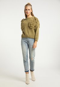 Olijfgroene gebreide trui met franjes op de schouders, gedragen met versleten lichtblauwe jeans en beige enkellaarzen.